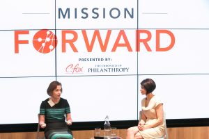 two woman on stage talking with the Mission Forward logo projected on a large screen behind them