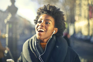 a Black woman in a winter coat and scarf with earbuds singing along and smiling
