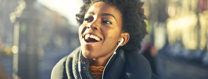 a Black woman in a winter coat and scarf with earbuds singing along and smiling