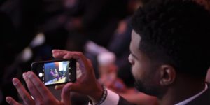A man holding a smart phone taking video of speakers onstage