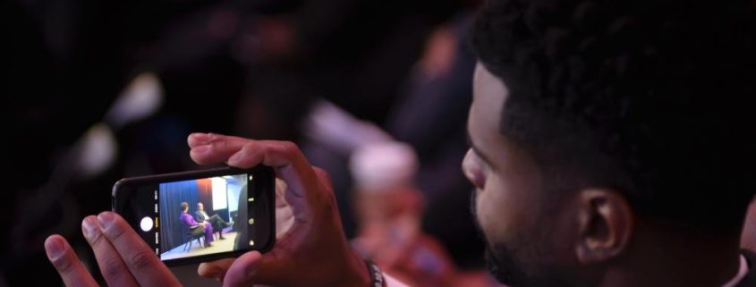 A man holding a smart phone taking video of speakers onstage