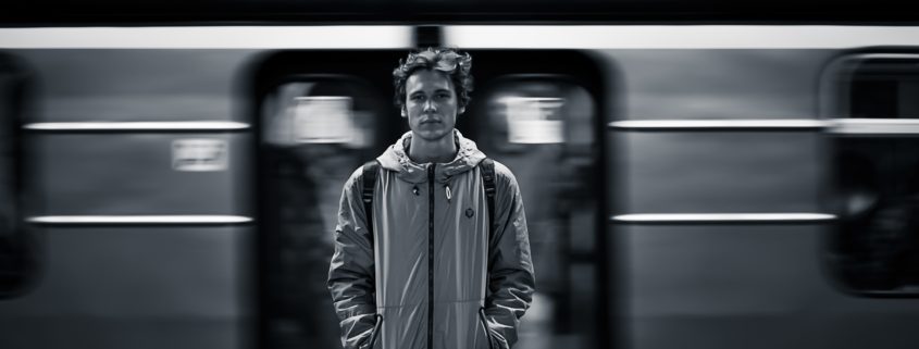 a young man looking directly at the camera as a subway train speeds behind him