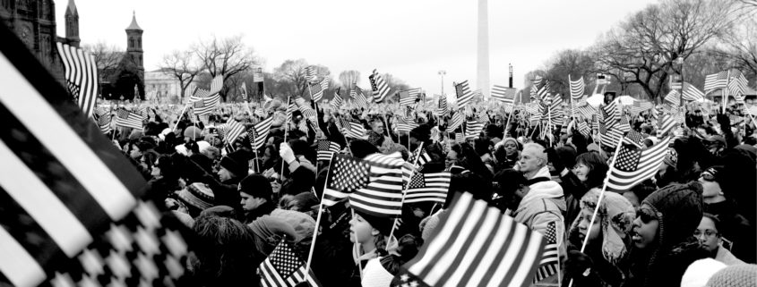 A crowd of people on the Mall in Washington, DC waving American flags. The Washington Monument is in the background.