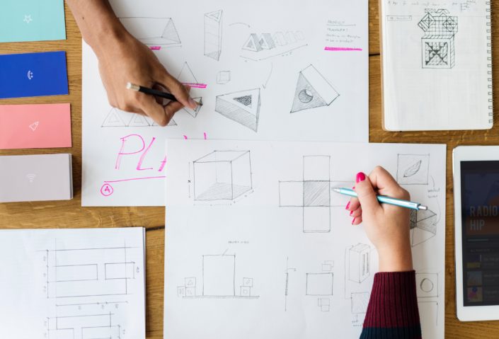 A top down view of sketches laid out on a table with two people working on them