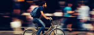A man in jeans and a t-shirt, with a backpack and headphones, riding a bicycle. The background of people behind him in blurred