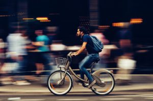 A man in jeans and a t-shirt, with a backpack and headphones, riding a bicycle. The background of people behind him in blurred
