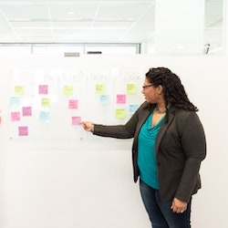 A woman in a green shirt stands at a board with many post it notes on it.