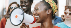 A young Black women wearing a colorful headband and classes, smiling, and holding a megaphone