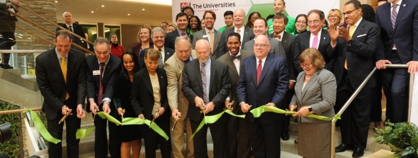 People dressed in suits, stand with scissors in front of a green ribbon. They are going to do a ribbon cutting ceremony