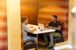 White man and black man working together in a booth. The black man has a laptop