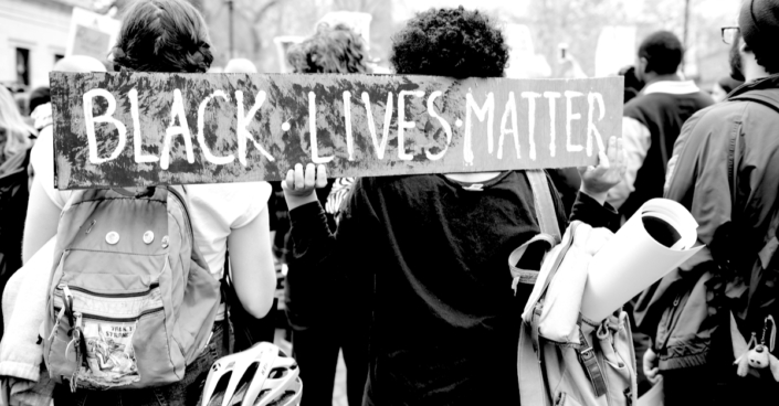 A black and white photo of protestors from behind holding a sign that says BLACK LIVES MATTER