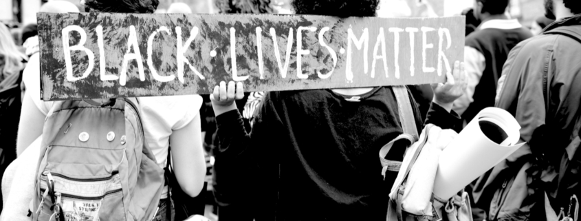 A black and white photo of protestors from behind holding a sign that says BLACK LIVES MATTER