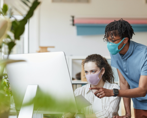 Portrait of two young coworkers wearing masks while pointing at computer screen in office