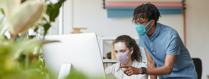 Portrait of two young coworkers wearing masks while pointing at computer screen in office