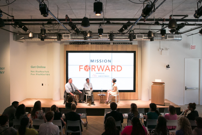 Audience in front of stage with a panel of 2 men being interviewed by a women.