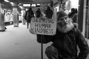 An older man with a beard and beanie on one knee holding a beat up sign that says "Seeking Human Kindness"