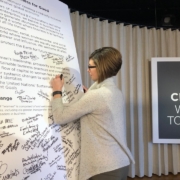 A woman signing an oversized document with a sign in the background that says We The Change, Women Together