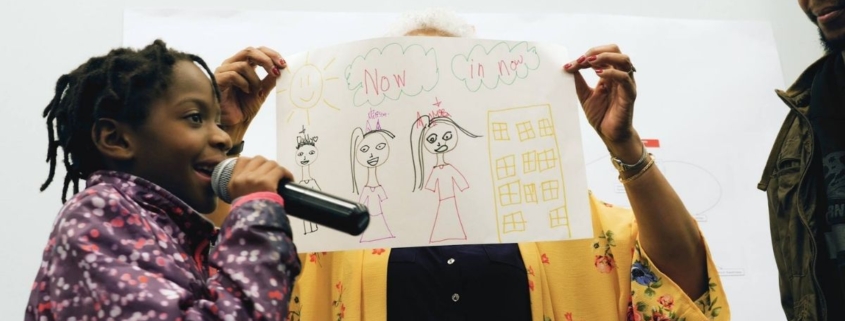 A girl stands with a sign and is on a microphone next to a sign that says now is now.