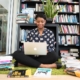 A black woman sits on the floor working on a laptop surrounded by books