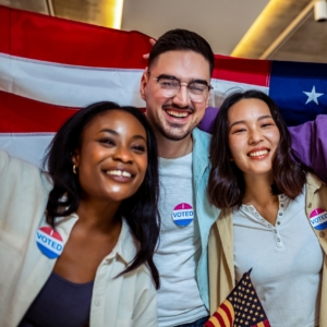 Three people with arms around each other, smiling, holding an American flag behind them, and wearing I Voted stickers.